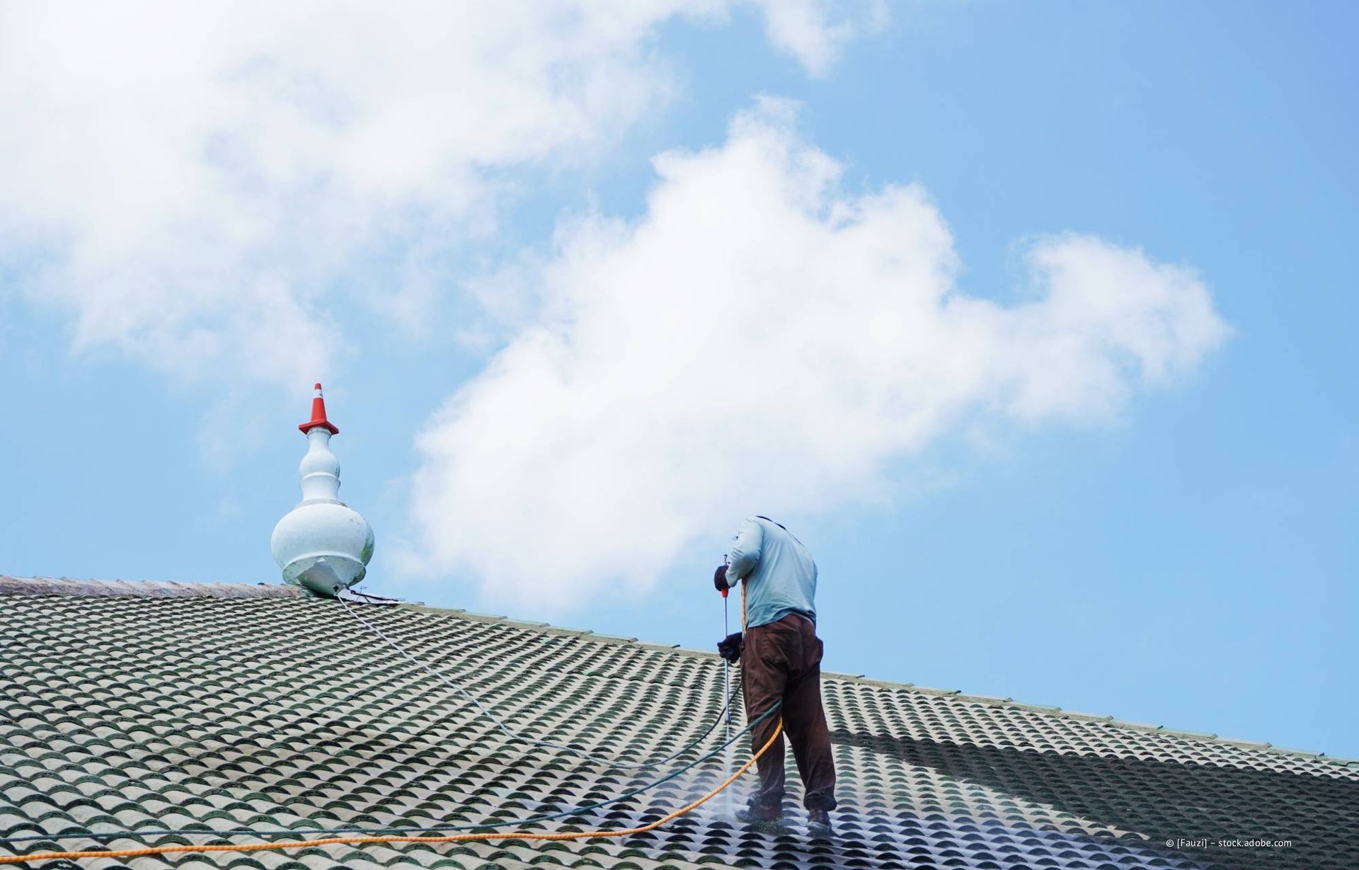 Eine Person reinigt ein grünes Ziegeldach mit einem Hochdruckreiniger; auf dem Dach steht ein verzierter Turm. Blauer Himmel mit Wolken.