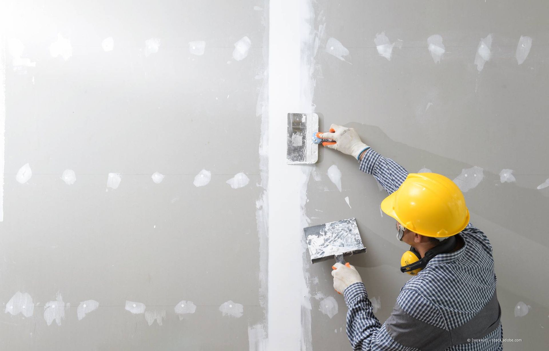 Eine Person mit gelbem Schutzhelm trägt mit einer Kelle Gipskartonspachtelmasse auf eine Wand auf.