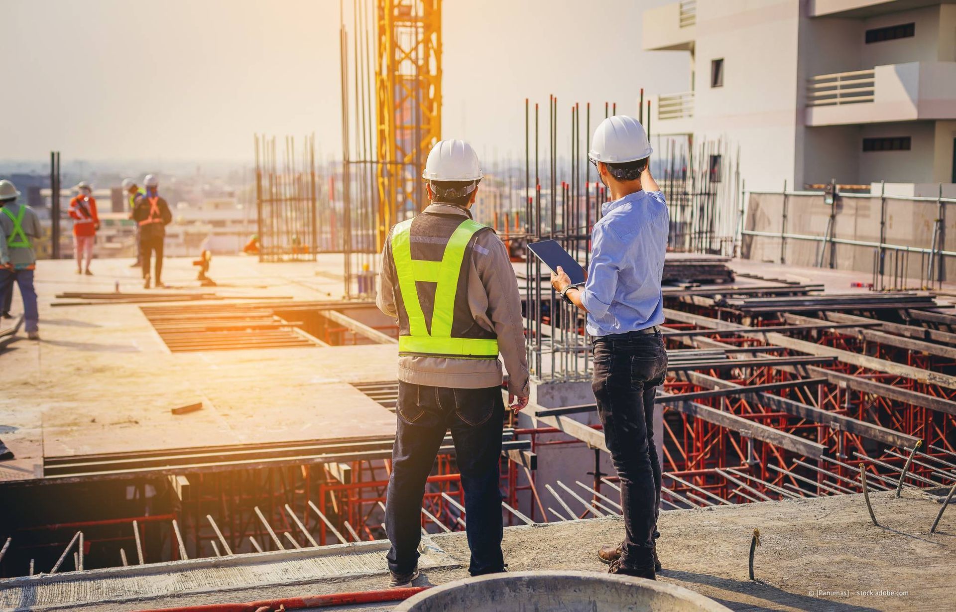 Bauarbeiter mit Schutzhelmen prüfen Baupläne eines im Bau befindlichen Gebäudes an einem sonnigen Tag.