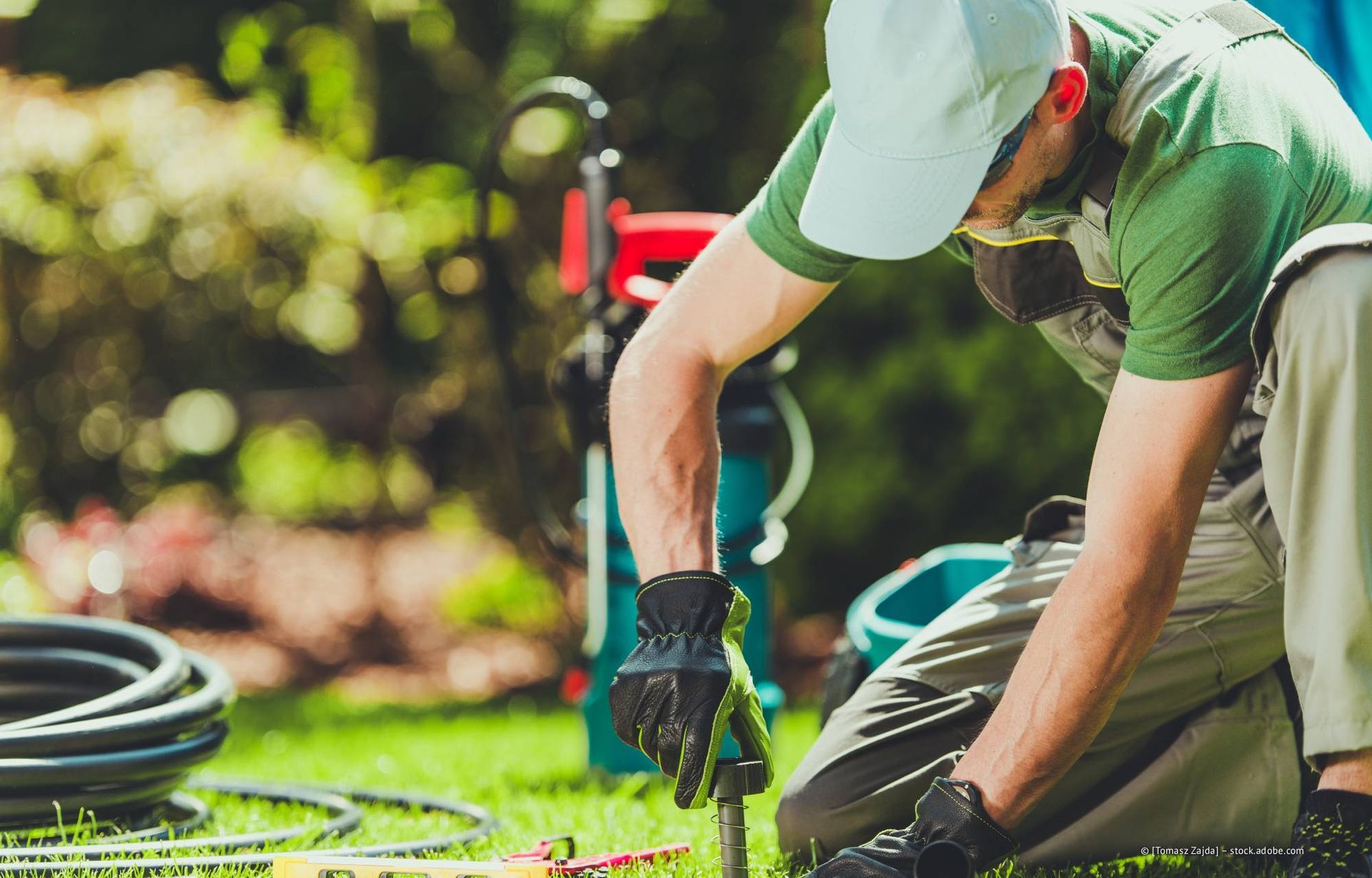 Ein Gärtner kniet im Freien und arbeitet an der Bewässerungsanlage. Er trägt eine Kappe, ein grünes Hemd und Handschuhe.