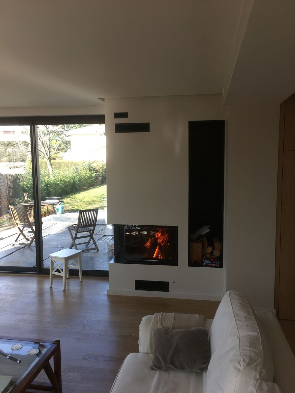 Salon moderne avec cheminée allumée, canapé blanc et portes-fenêtres ouvertes sur un jardin.