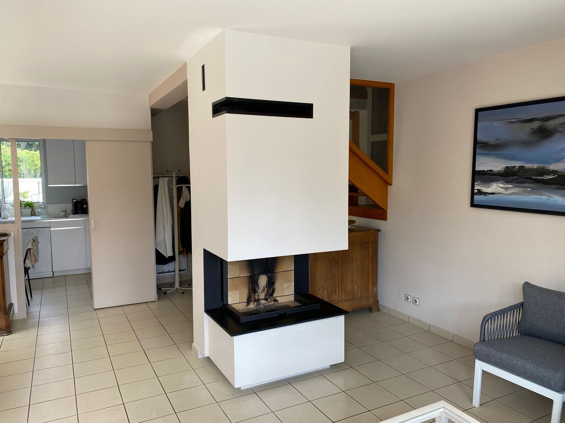 Salon lumineux avec cheminée blanche, escalier et cuisine visibles en arrière-plan.