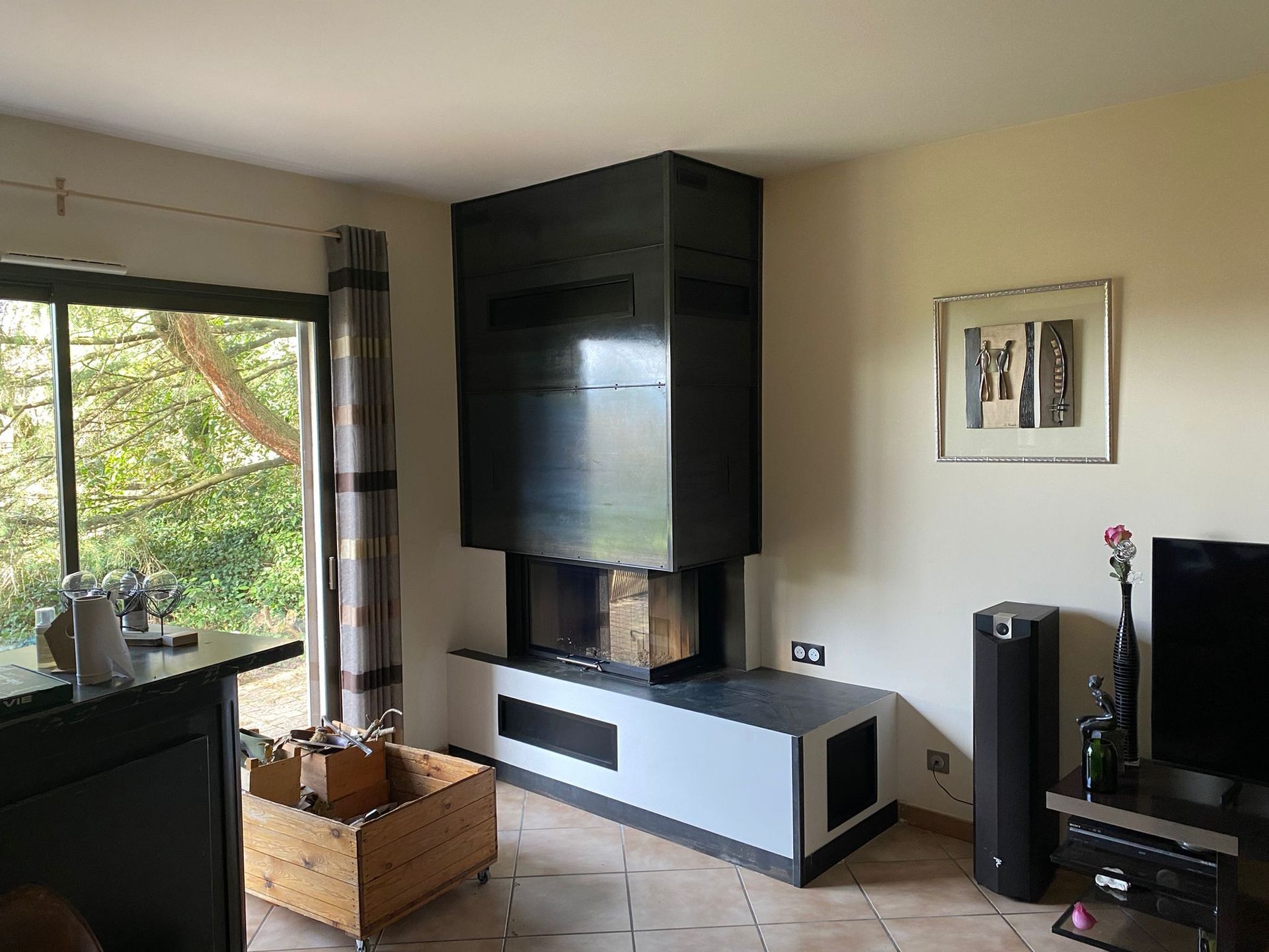 Salon moderne avec cheminée noire, télévision, grande fenêtre et panier en osier contre un mur beige.