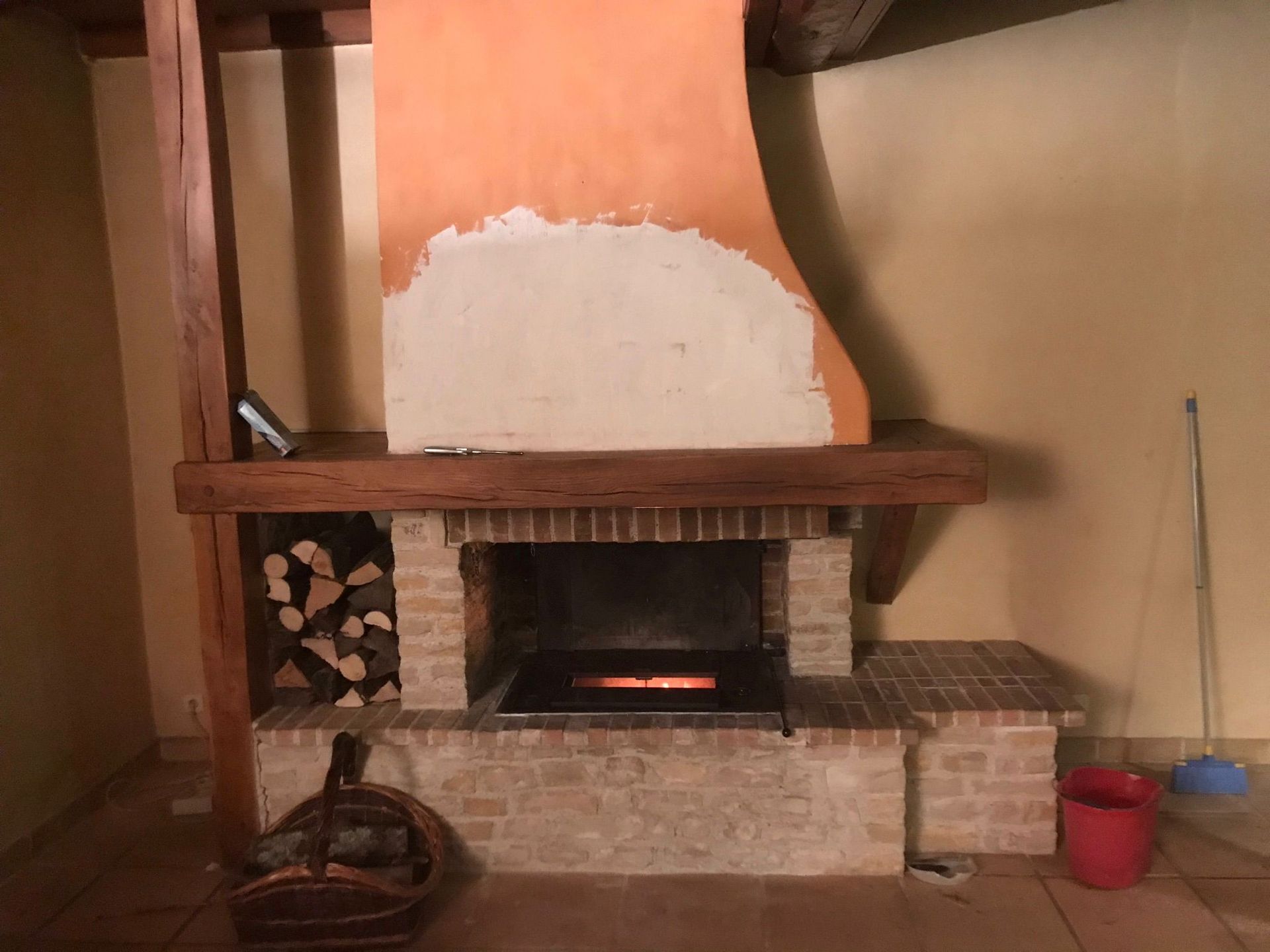 Cheminée en pierre avec une grande hotte orange, une étagère en bois et un seau rouge à côté.