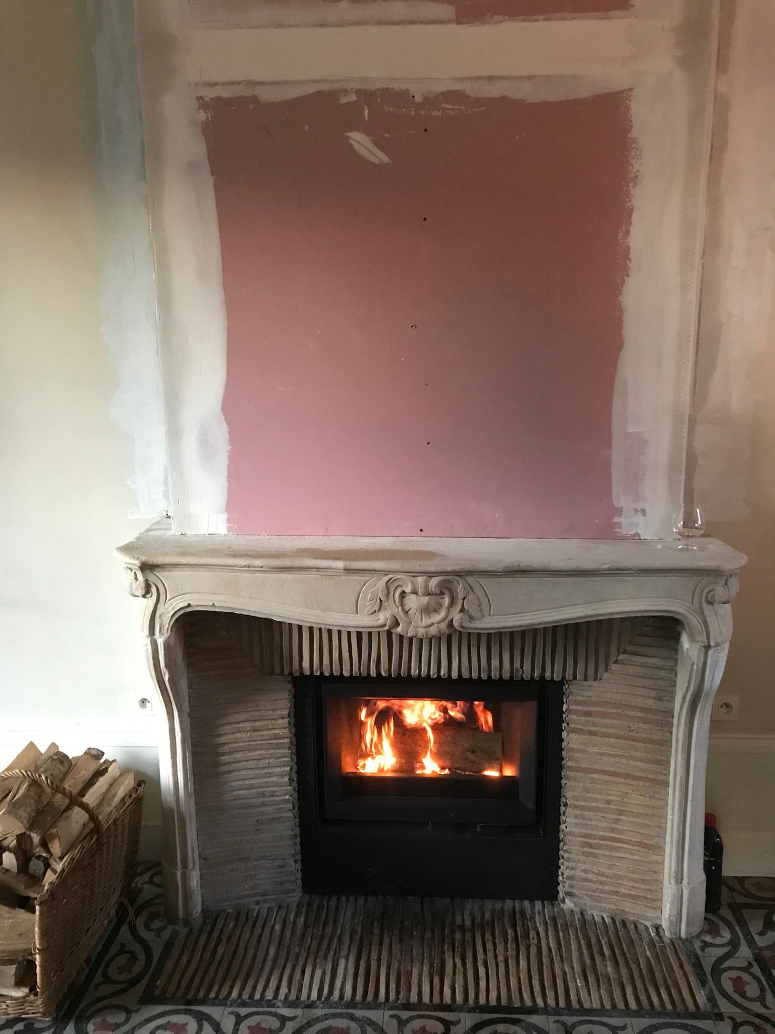 Un feu brûle dans une cheminée noire avec un manteau blanc et un mur rose inachevé au-dessus.