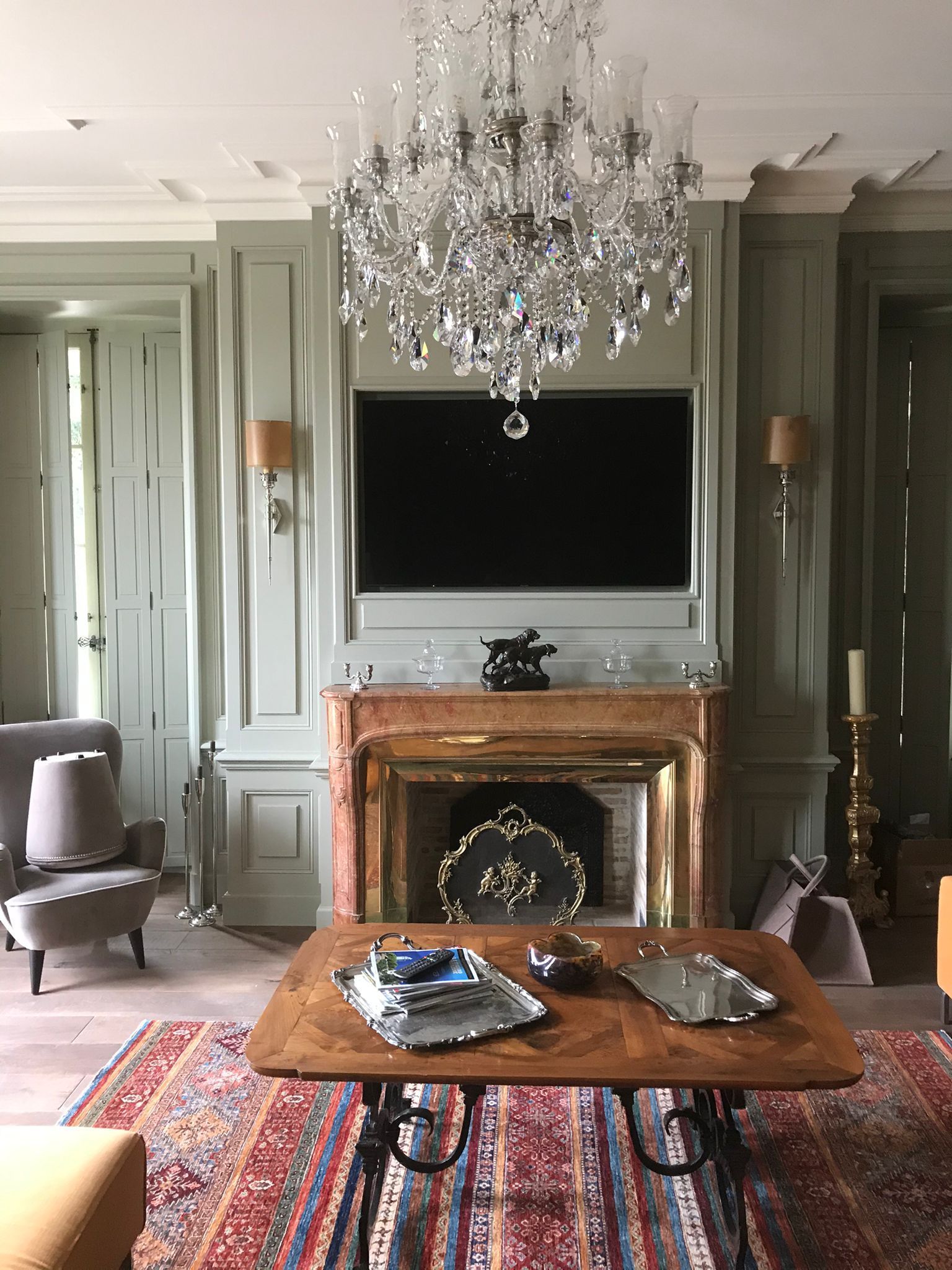 Salon élégant avec lustre en cristal, cheminée, grand miroir et table basse en bois sur un tapis à motifs.