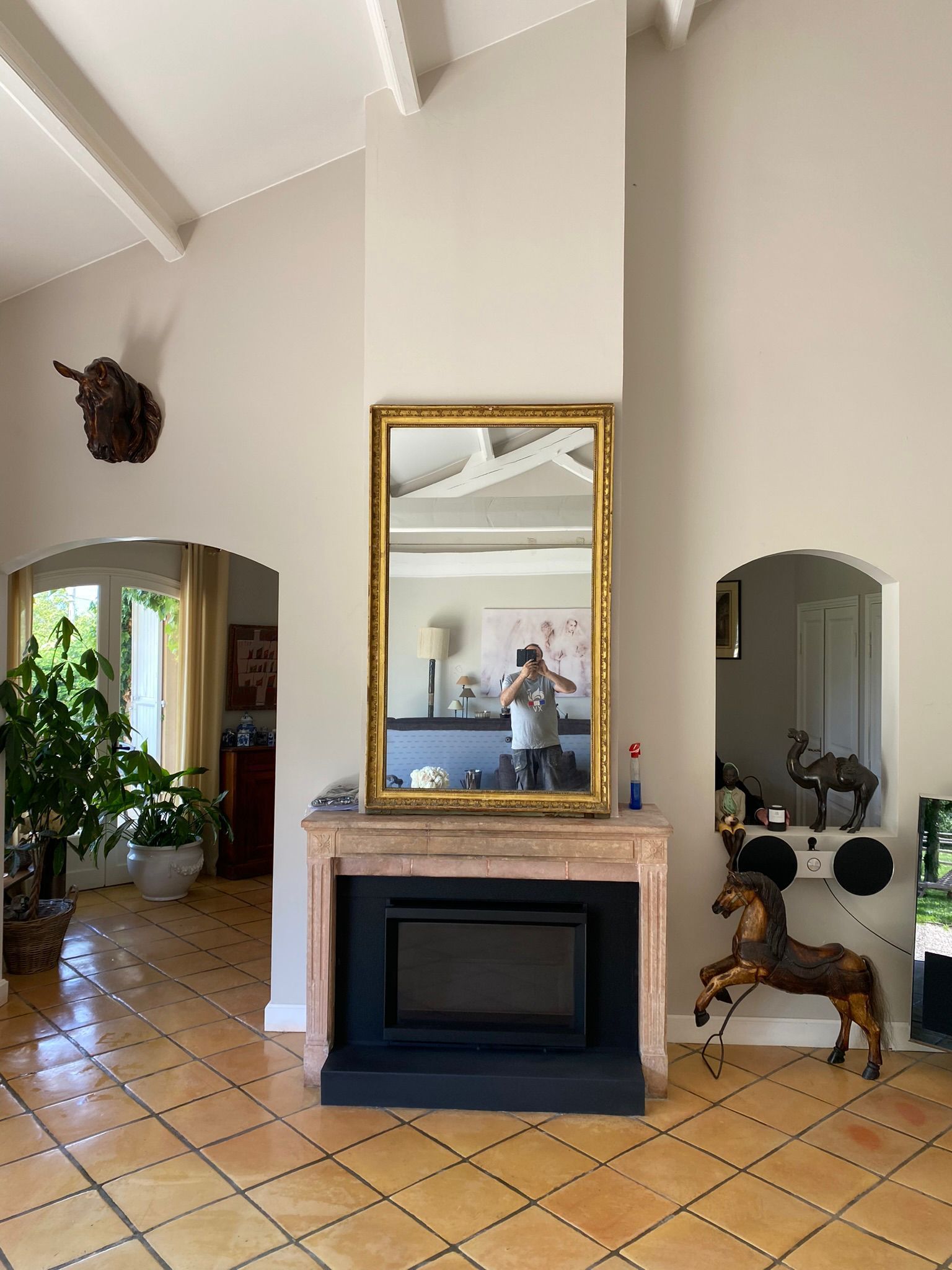 Entrée lumineuse avec portes cintrées, cheminée, grand miroir doré et sol carrelé agrémenté de plantes et d'éléments décoratifs.