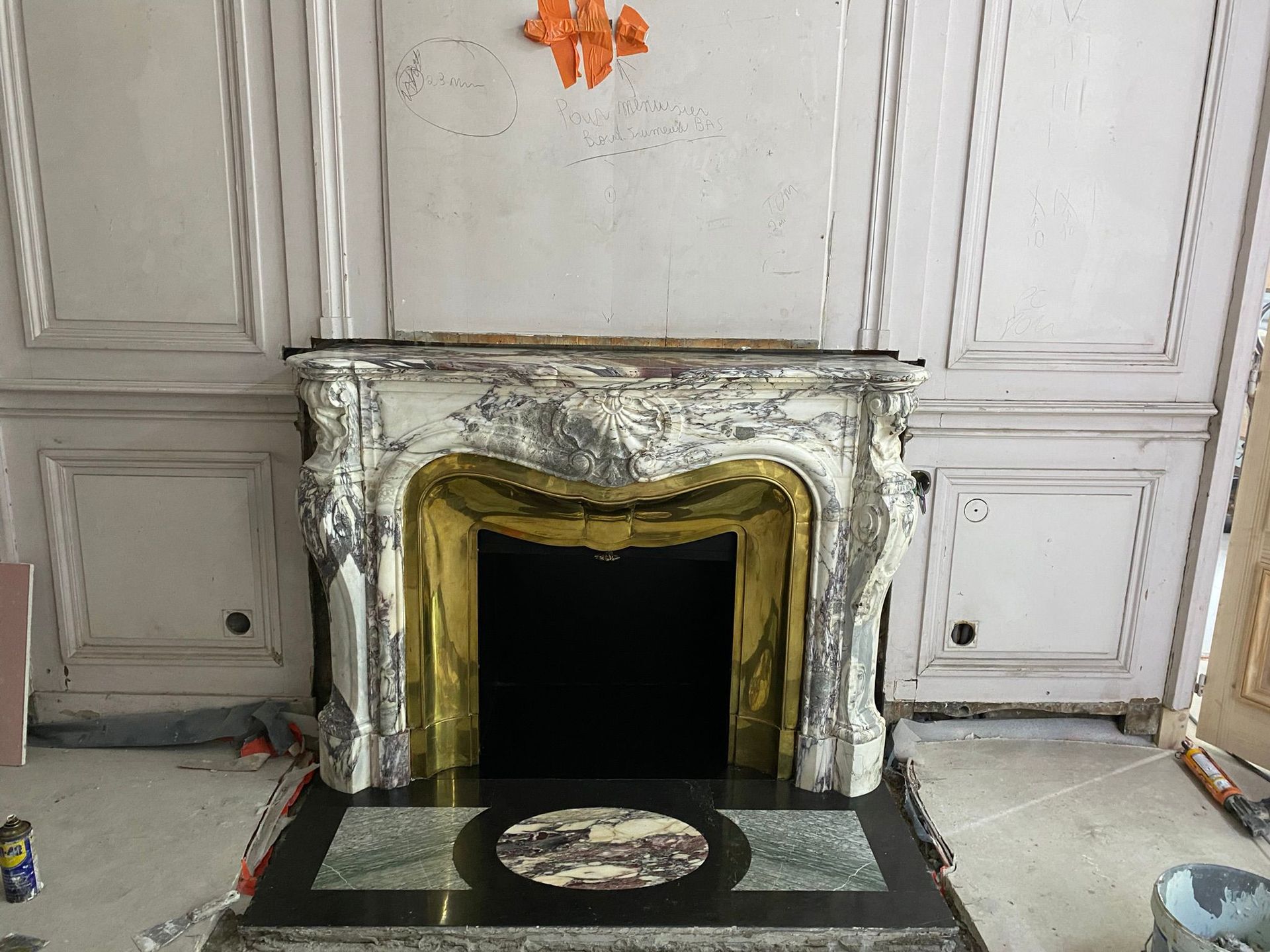 Cheminée en marbre blanc ornée de garnitures dorées dans une pièce lambrissée, foyer sombre et vide et âtre noir.