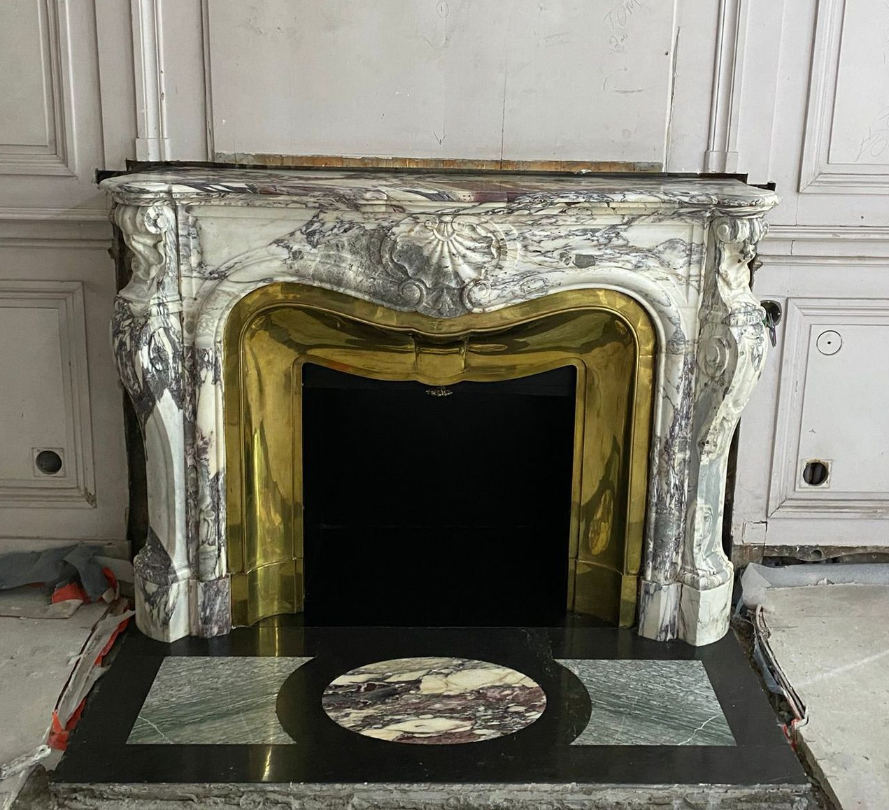 Salon élégant avec lustre en cristal, cheminée, grand miroir et table basse en bois sur un tapis à motifs.