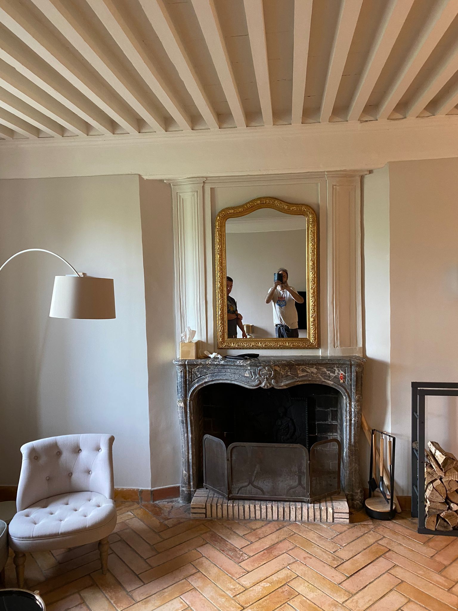 Salon élégant avec plafond à rayures, cheminée ornée, miroir et fauteuil capitonné sur parquet à chevrons.