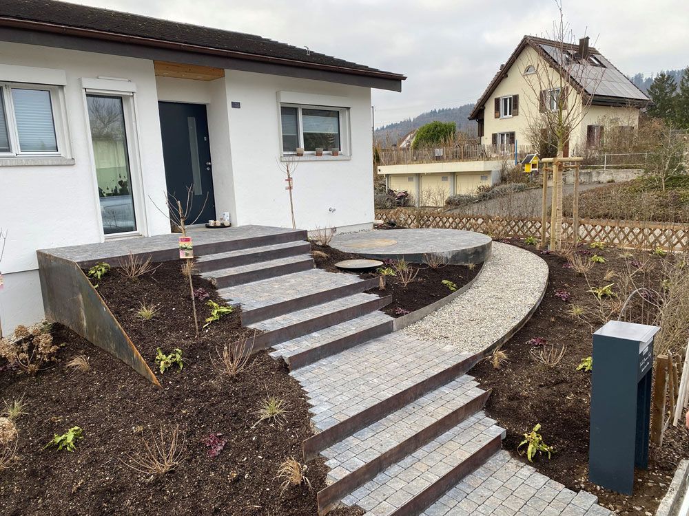 Steinstufen führen zu einem Hauseingang, der von dunkler Erde und Gartenanlagen umgeben ist. Auf dem Treppenabsatz steht ein Rasenmäher.