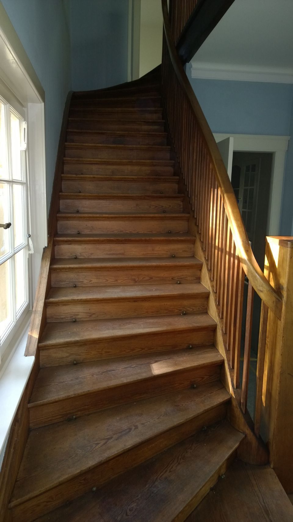 Holztreppe mit Geländer in einem blau gestrichenen Innenflur, von links durch Tageslicht beleuchtet.