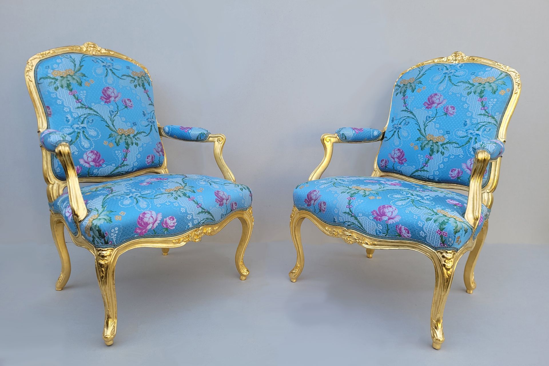 Zwei kunstvoll verzierte blaue Sessel mit floralem Muster und goldenen Gestellen vor grauem Hintergrund