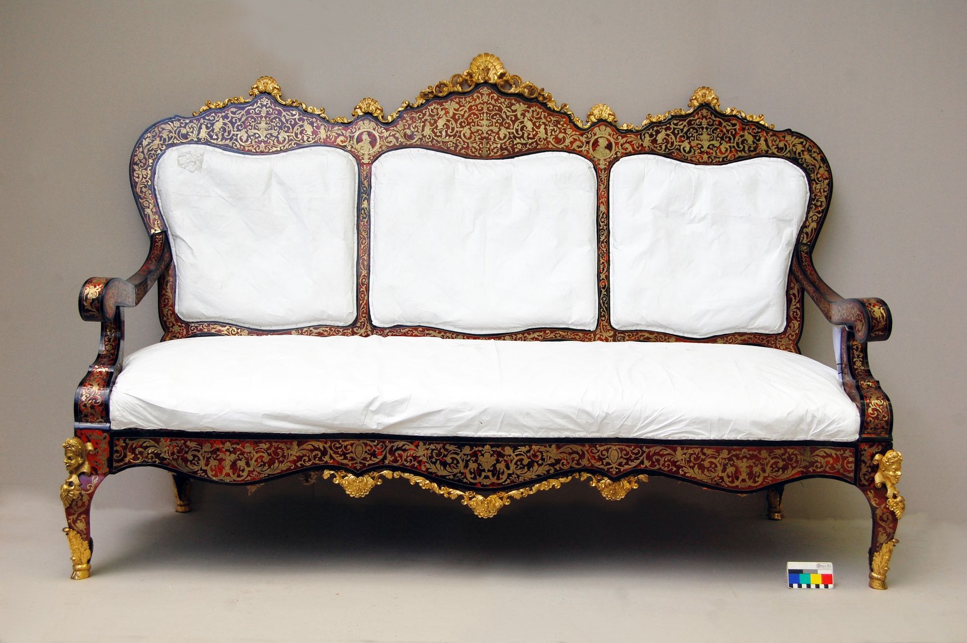 Verschnörkeltes Vintage-Sofa aus Holz mit weißen Kissen und goldenen Akzenten vor einem schlichten Hintergrund