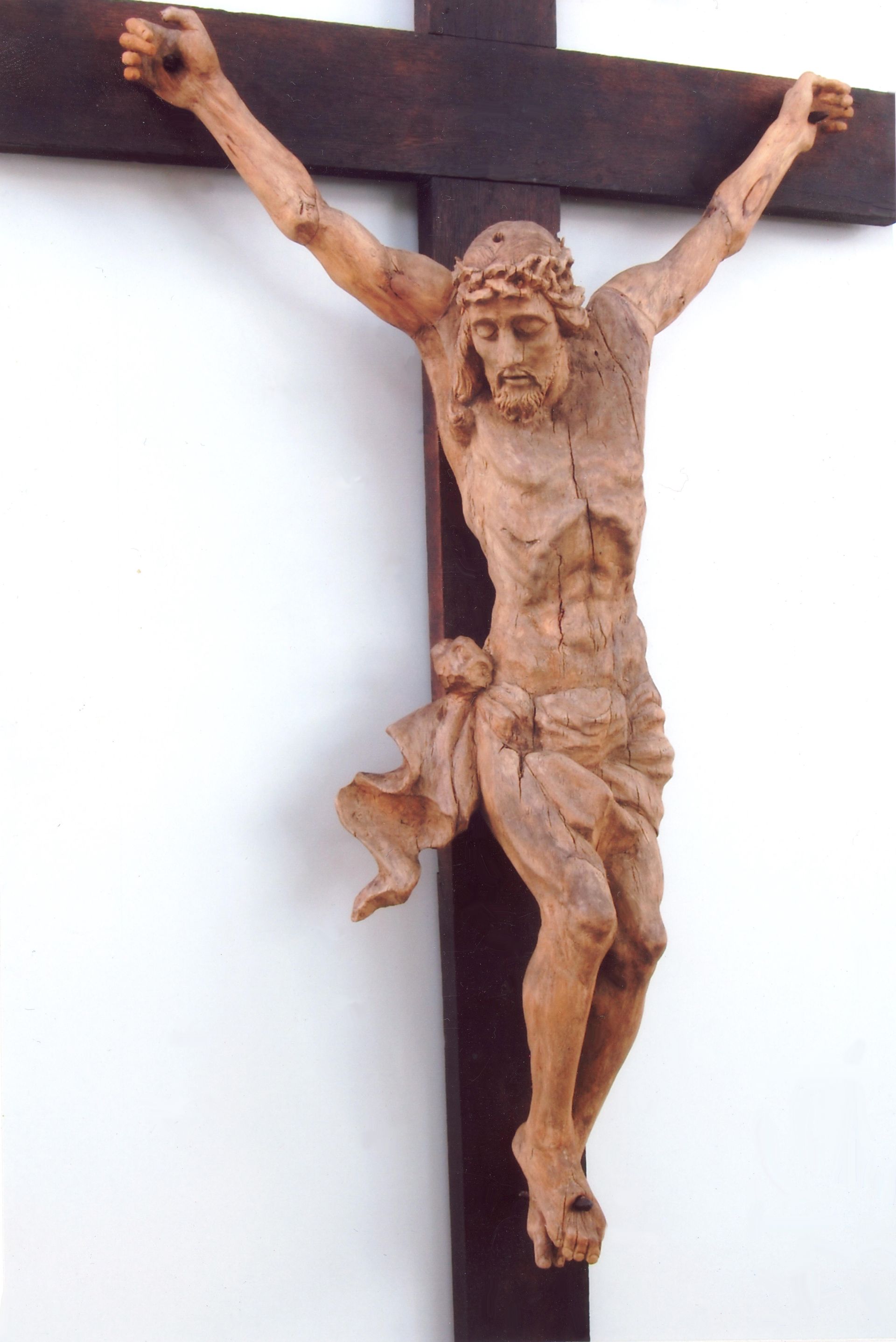 Holzskulptur eines gekreuzigten Jesus mit ausgebreiteten Armen an einem dunklen Kreuz vor einer weißen Wand