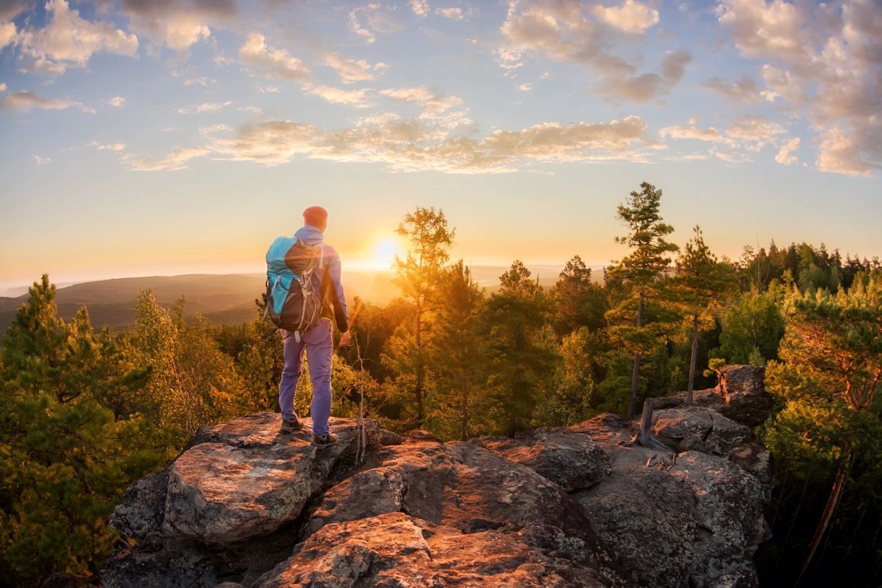 a man with a backpack is standing on top of a rocky cliff overlooking a forest at sunset .