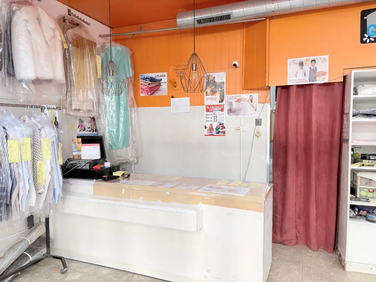 El interior de una tintorería con un mostrador blanco, ropa colgada en tendederos de plástico y paredes.