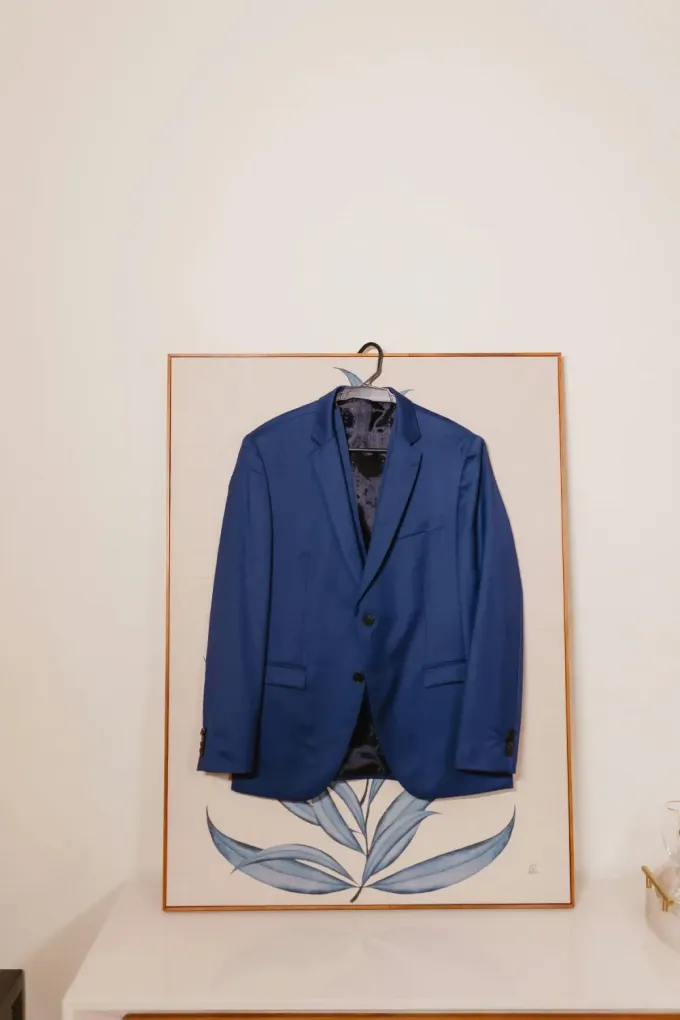 Una chaqueta de traje azul colgada en una percha sobre una lámina enmarcada de hojas azules sobre una pared blanca.