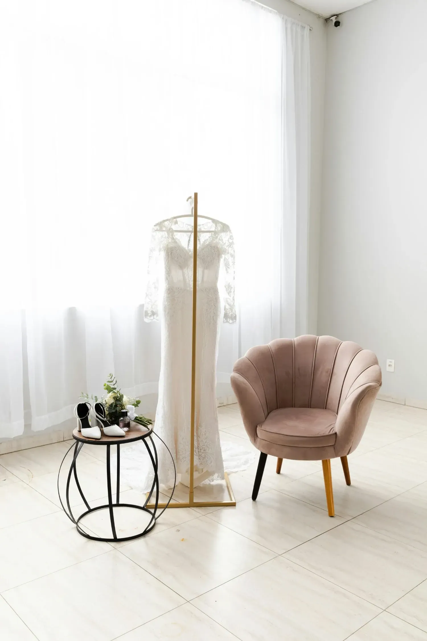 Un vestido de novia blanco cuelga de un soporte dorado junto a una mesa auxiliar redonda con accesorios.