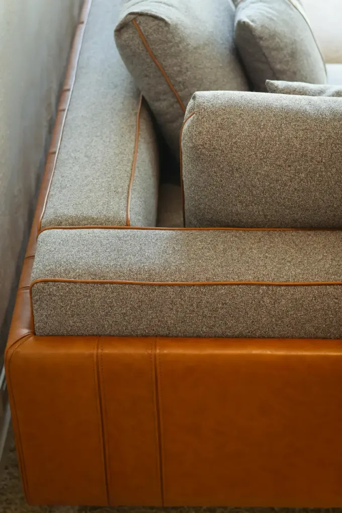 Primer plano de un sofá moderno con estructura de cuero marrón y cojines de tela texturizada de color gris claro.