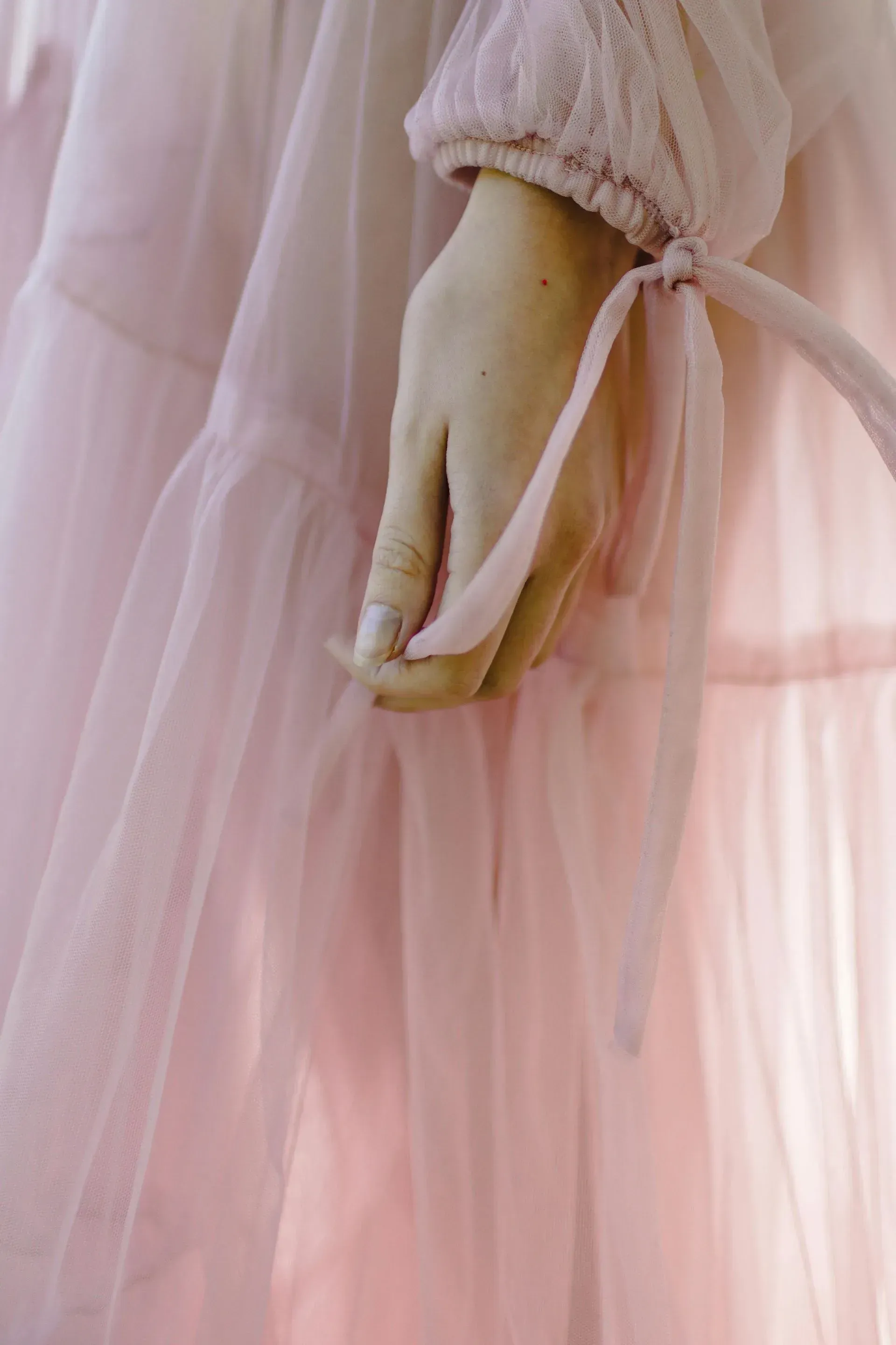 Primer plano de una mano que sostiene con delicadeza la tela rosa fluida y la cinta de un vestido de tul escalonado.
