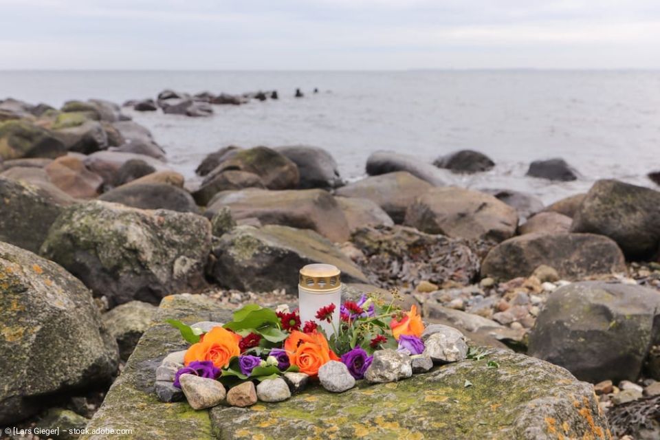 Eine Gedenkkerze und bunte Blumen wurden unter bedecktem Himmel an einem felsigen Ufer am Meer aufgestellt.