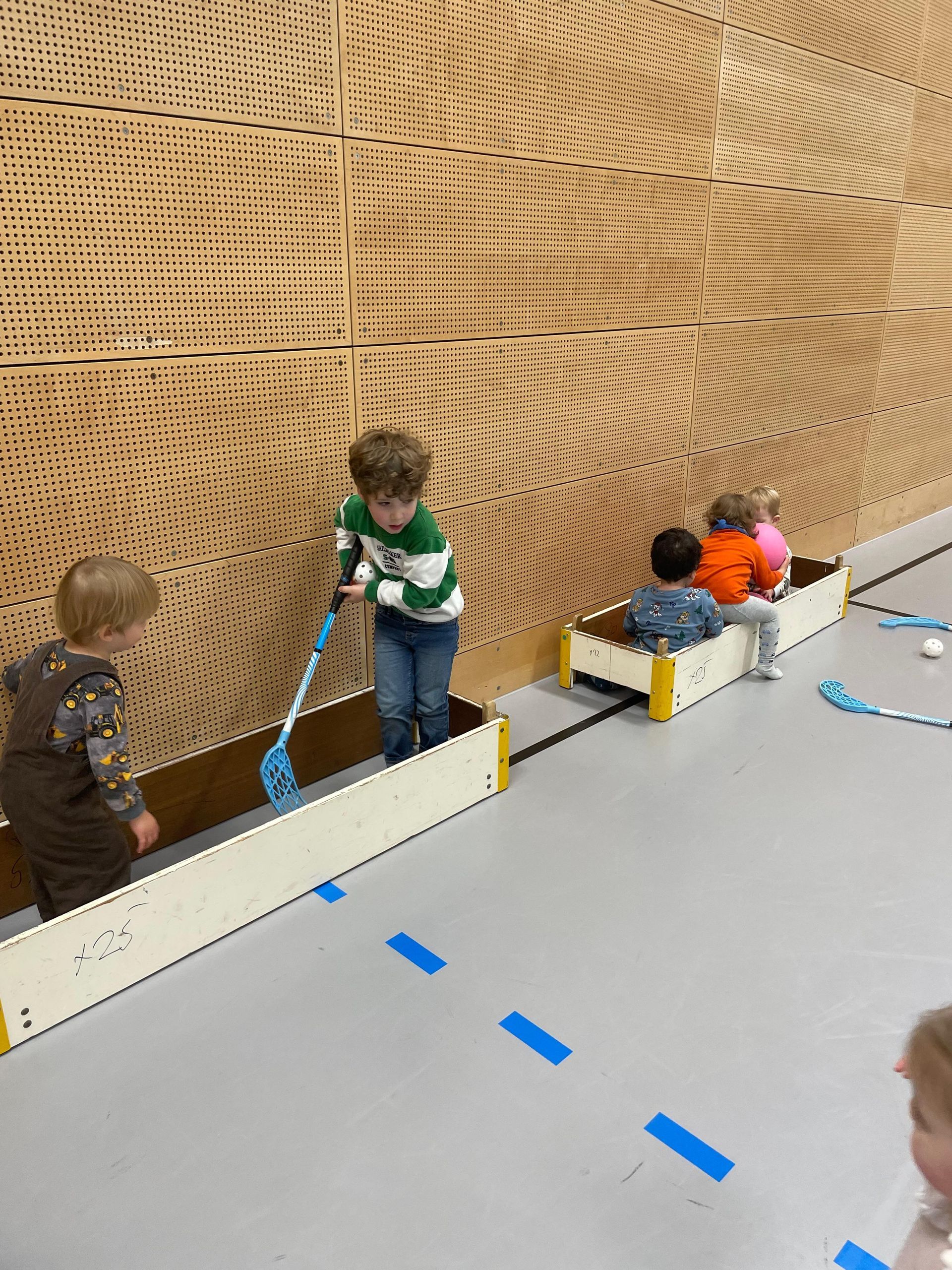 Kinder spielen Unihockey in einer Turnhalle. Ein Kind schwingt einen Hockeyschläger, andere sitzen in Holzkisten.