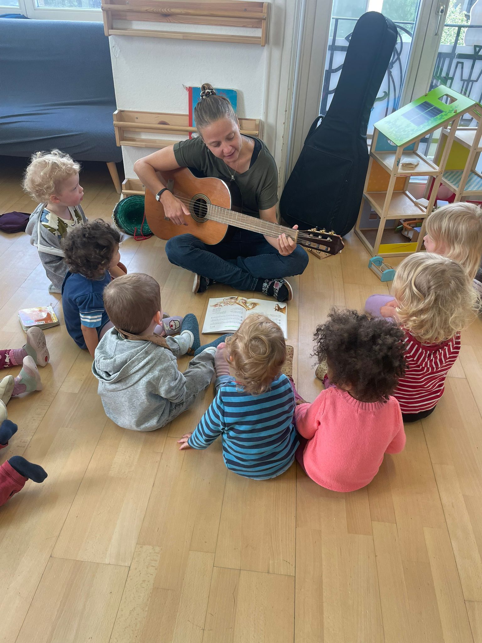 Eine Frau spielt Gitarre, umgeben von Kindern, die drinnen im Kreis sitzen.