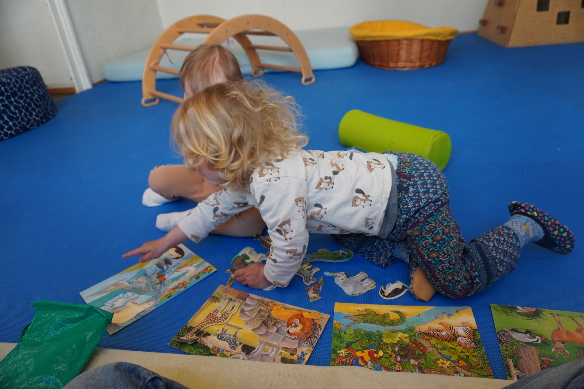 Kind auf blauer Matte, spielt mit Puzzleteilen, ein weiteres Kind im Hintergrund.