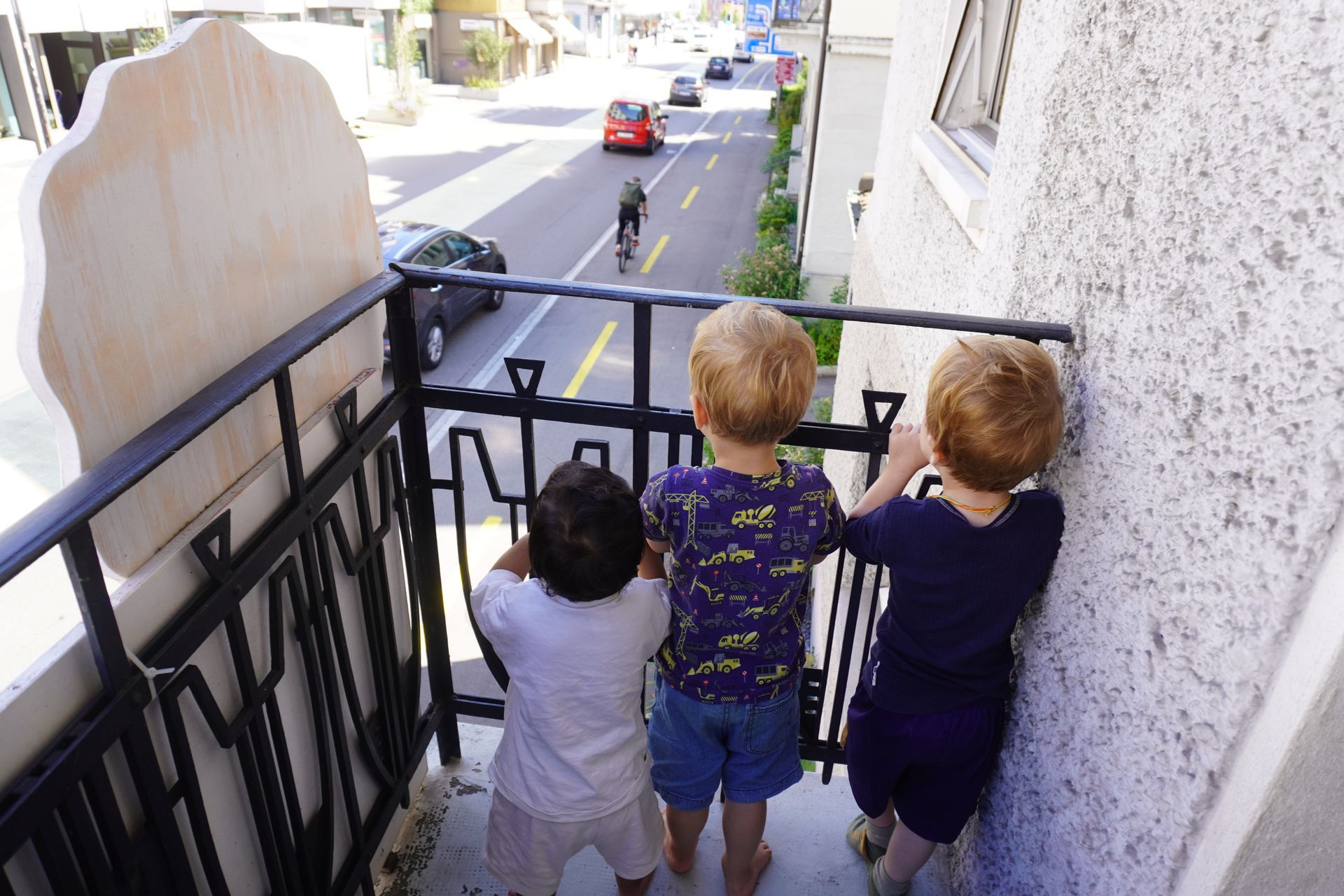 Drei Kinder stehen auf einem Balkon und schauen auf eine Straße mit Autos und einem Radfahrer.