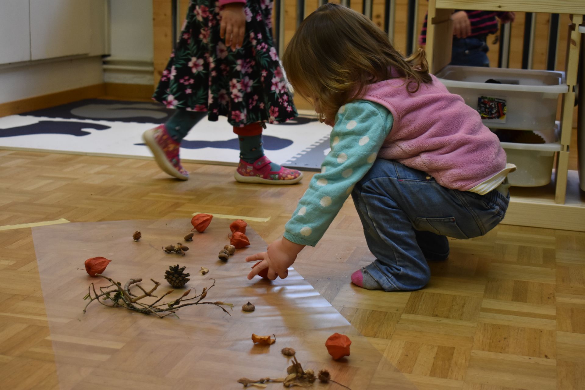 Ein kniendes Kind spielt mit Naturgegenständen auf dem Boden, ein anderes Kind geht vorbei.