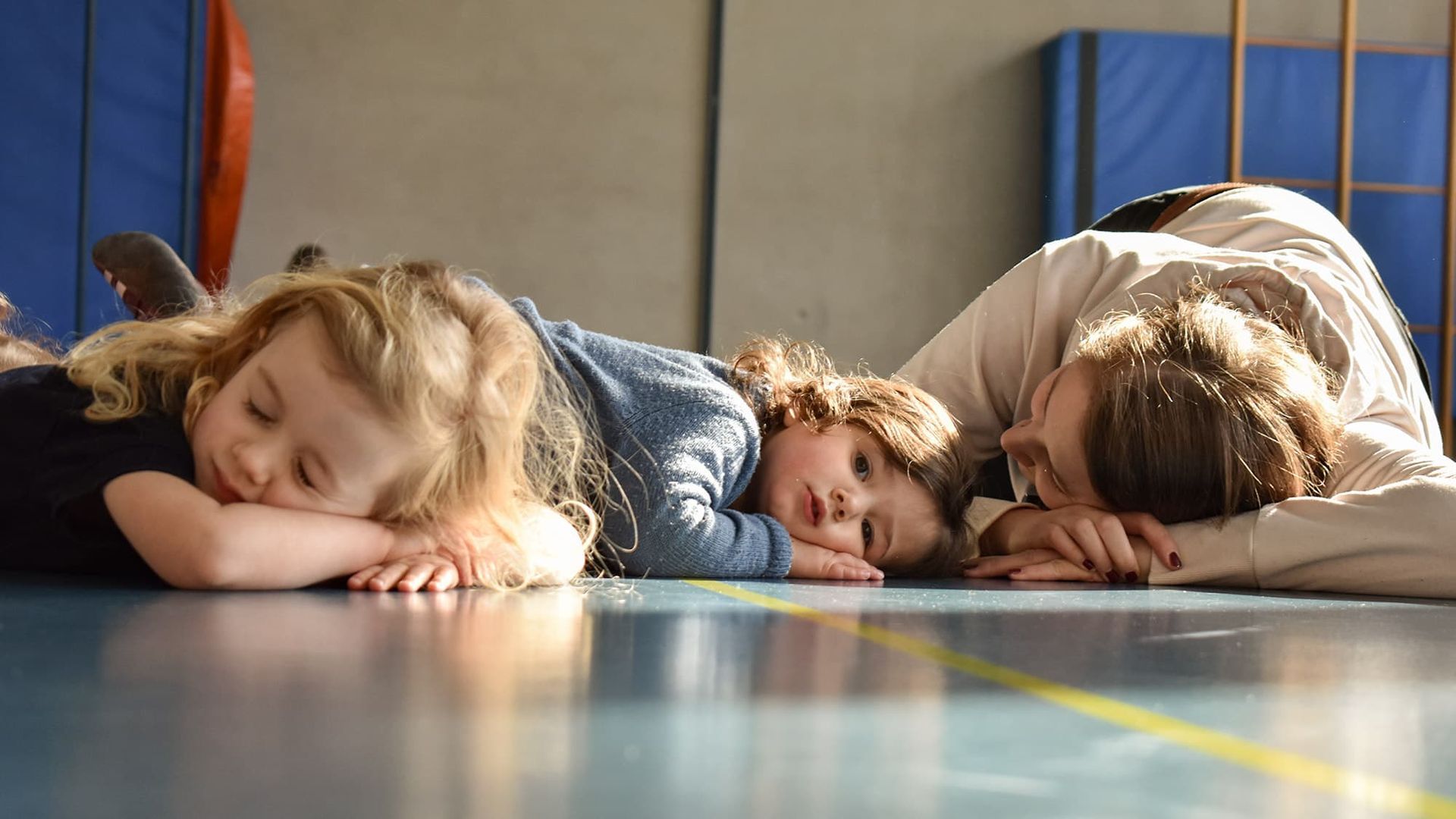 Drei Personen liegen mit gesenkten Köpfen auf dem Boden einer Turnhalle. 