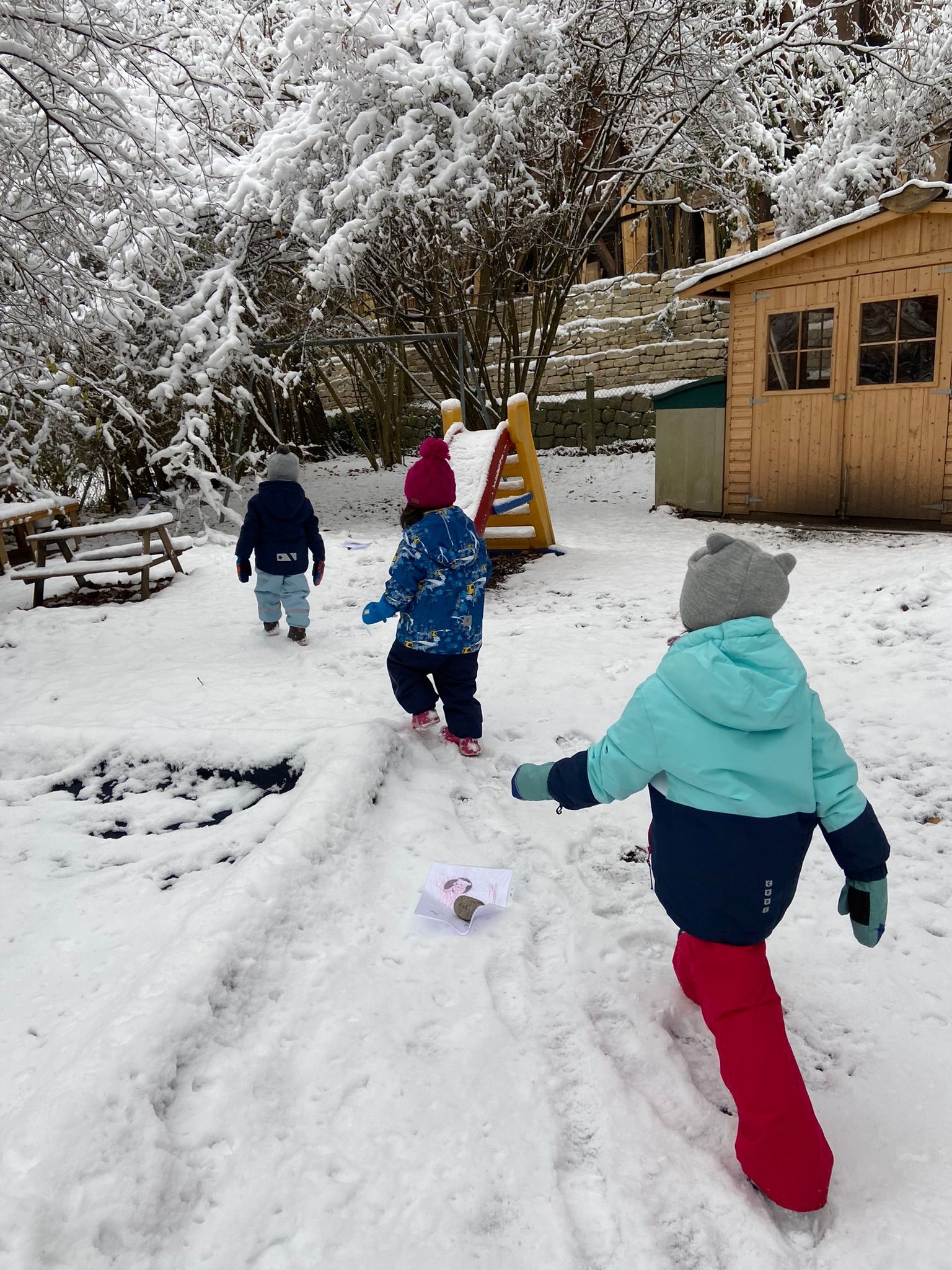 Kinder spielen im Schnee auf einem verschneiten Hof in der Nähe eines hölzernen Spielhauses und Bäumen.