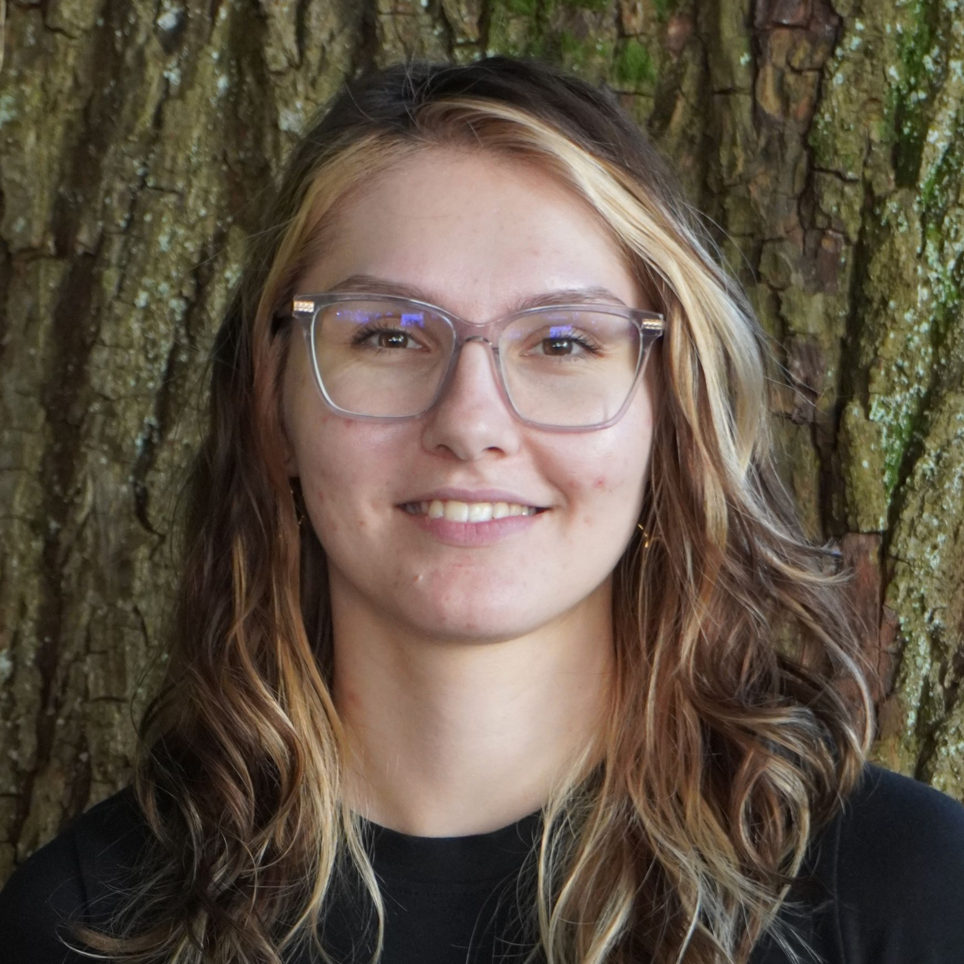 Ein junger Mensch mit Brille lächelt vor einem strukturierten Baumstamm.