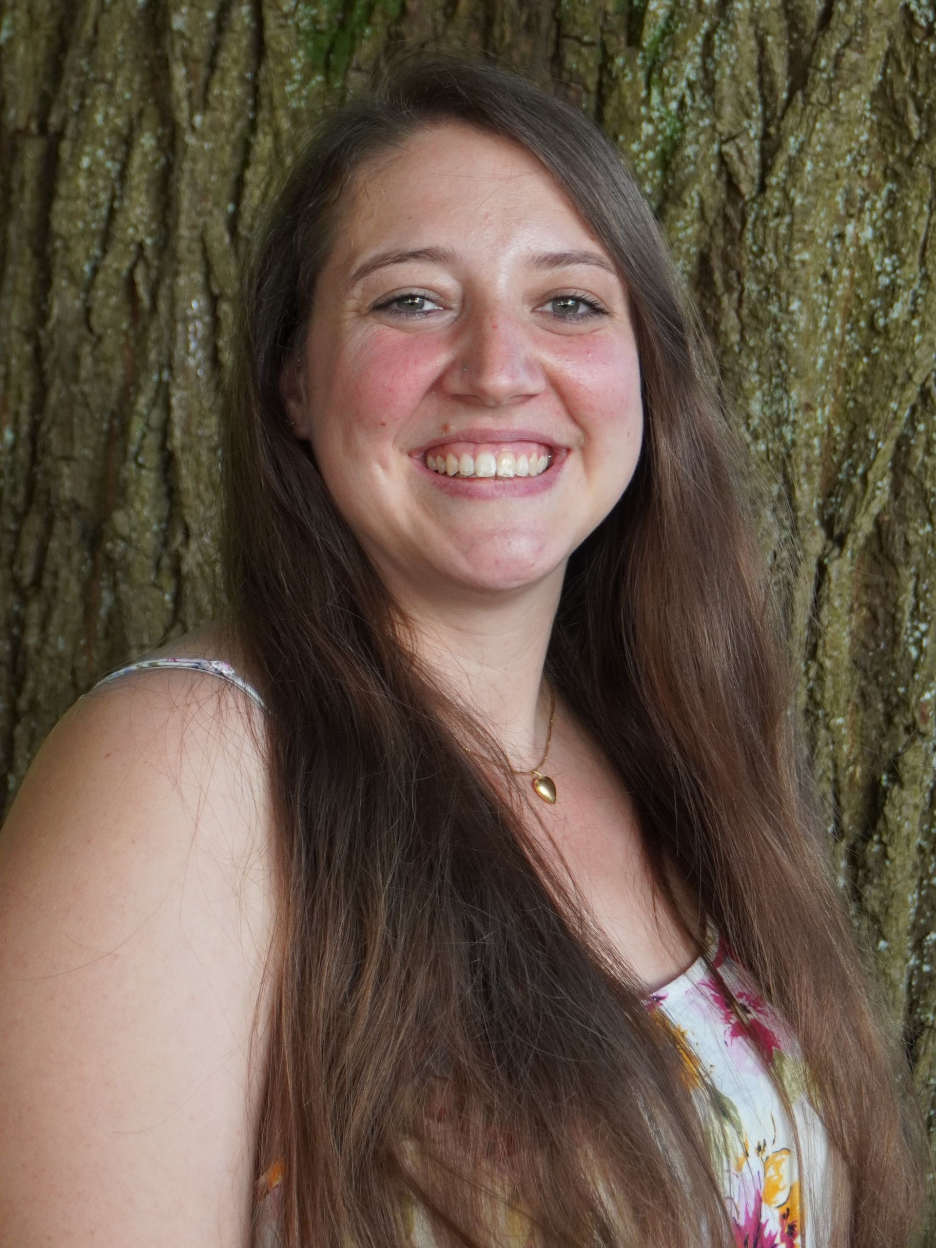 Eine lächelnde Frau mit langen braunen Haaren, die ein weißes Oberteil mit Blumenmuster trägt, steht vor einem Baum.