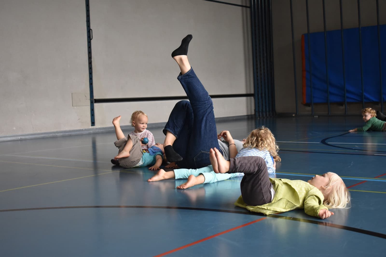 Szene in einer Turnhalle. Ein Erwachsener mit hochgelegtem Bein ist von kleinen Kindern umgeben.