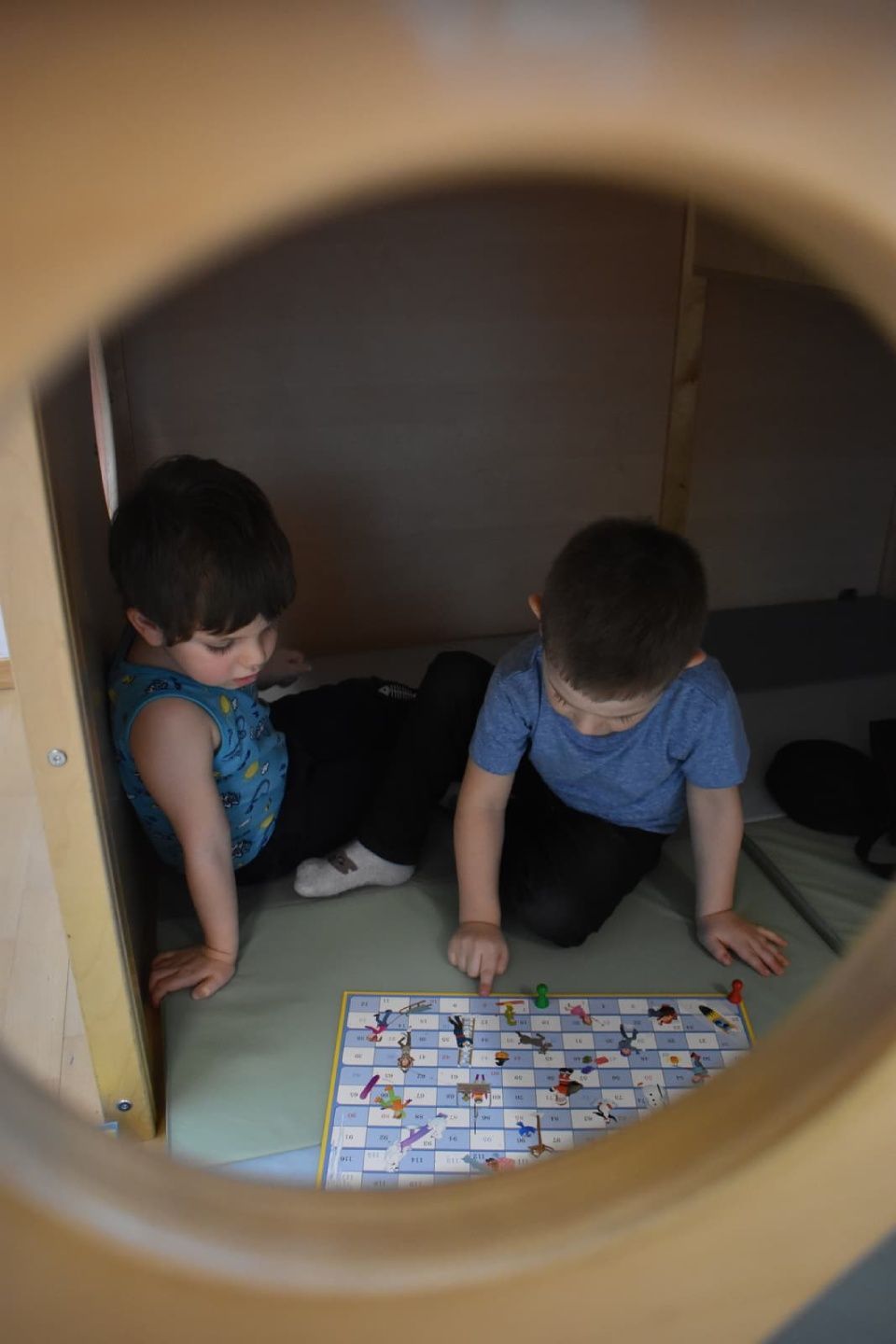 Zwei kleine Kinder spielen in einem Holzbauwerk ein Brettspiel; eines greift nach einem Spielstein.