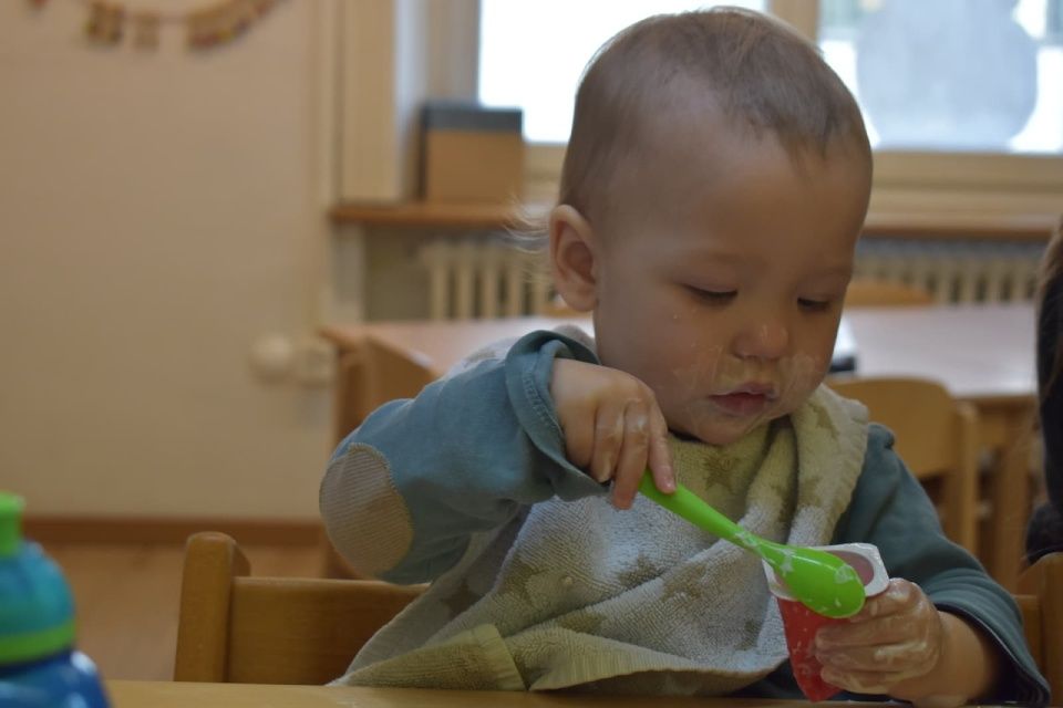 Kind mit verschmiertem Gesicht, das am Tisch einen grünen Löffel hält.