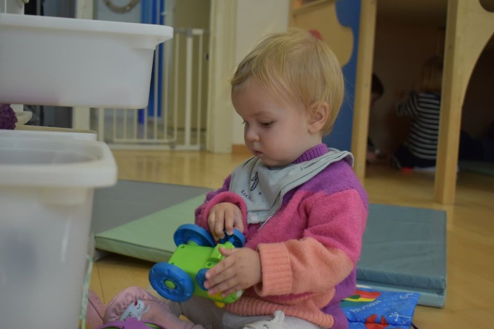 Kleinkind spielt drinnen mit einem grünen Spielzeugauto, trägt einen rosa Pullover und ein Lätzchen und ist konzentriert.