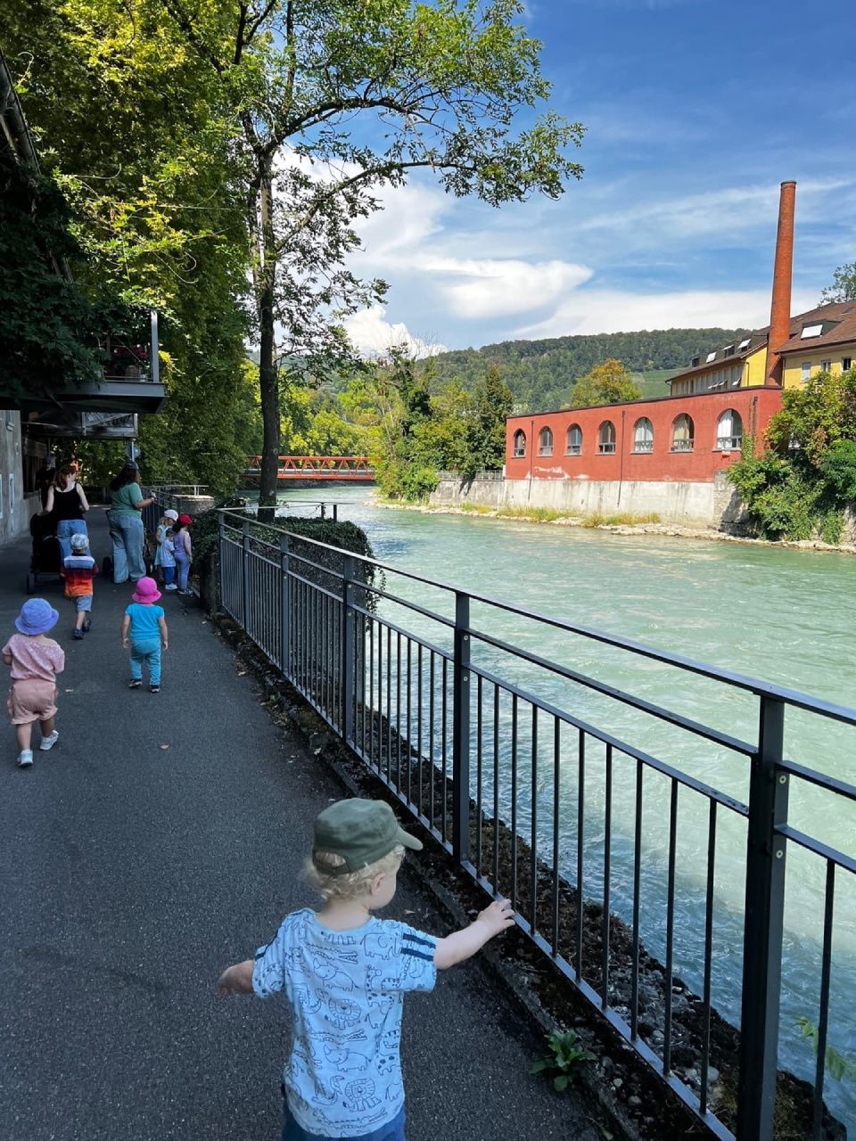 Kinder spazieren einen Uferweg entlang und passieren dabei ein Backsteingebäude mit roter Fassade und rotem Schornstein.