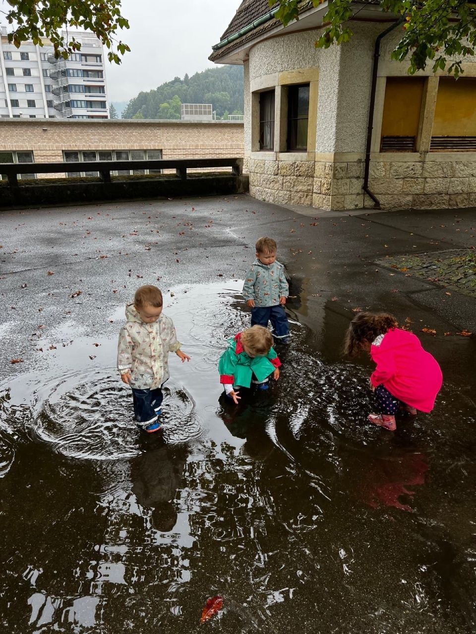 Vier kleine Kinder spielen in einer großen Pfütze auf einem nassen, gepflasterten Untergrund.