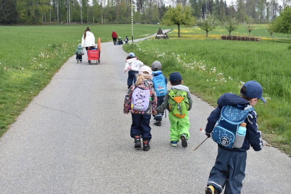 Kinder mit Rucksäcken gehen einen Weg entlang, gefolgt von Erwachsenen und einem Kinderwagen in einem Park.