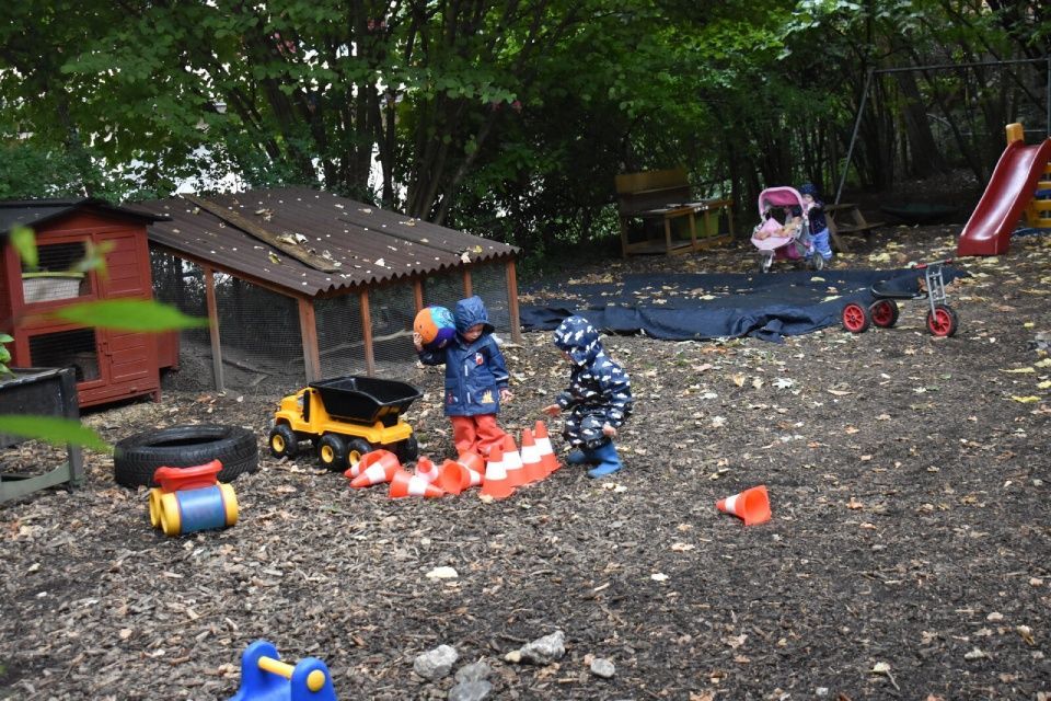 Kinder spielen mit Spielzeug und Hütchen auf einem bewaldeten Spielplatz in der Nähe eines Hühnerstalls und einer Rutsche.