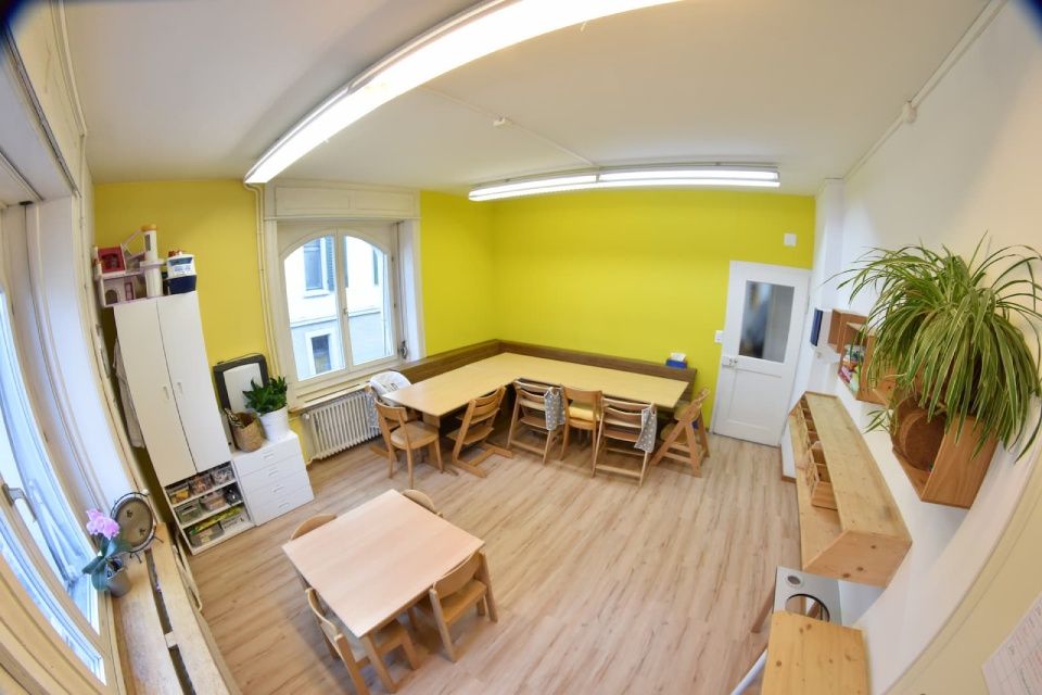 Kinderfreundliches Zimmer mit Tischen und Stühlen; gelbe Wände und ein großes Fenster.