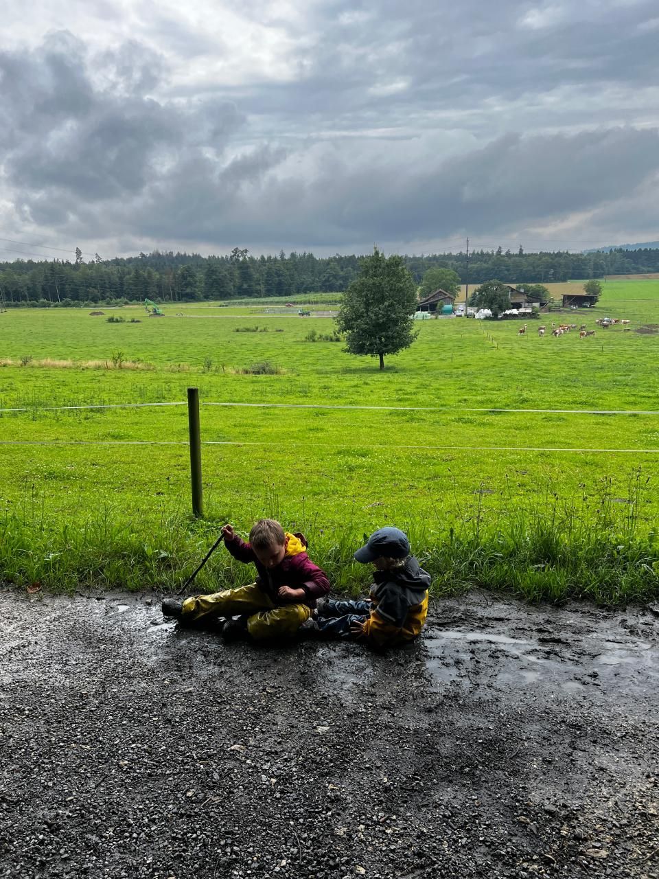 Zwei Kinder spielen im Schlamm auf einer nassen Straße vor einem grünen Feld. Bewölkter Himmel.