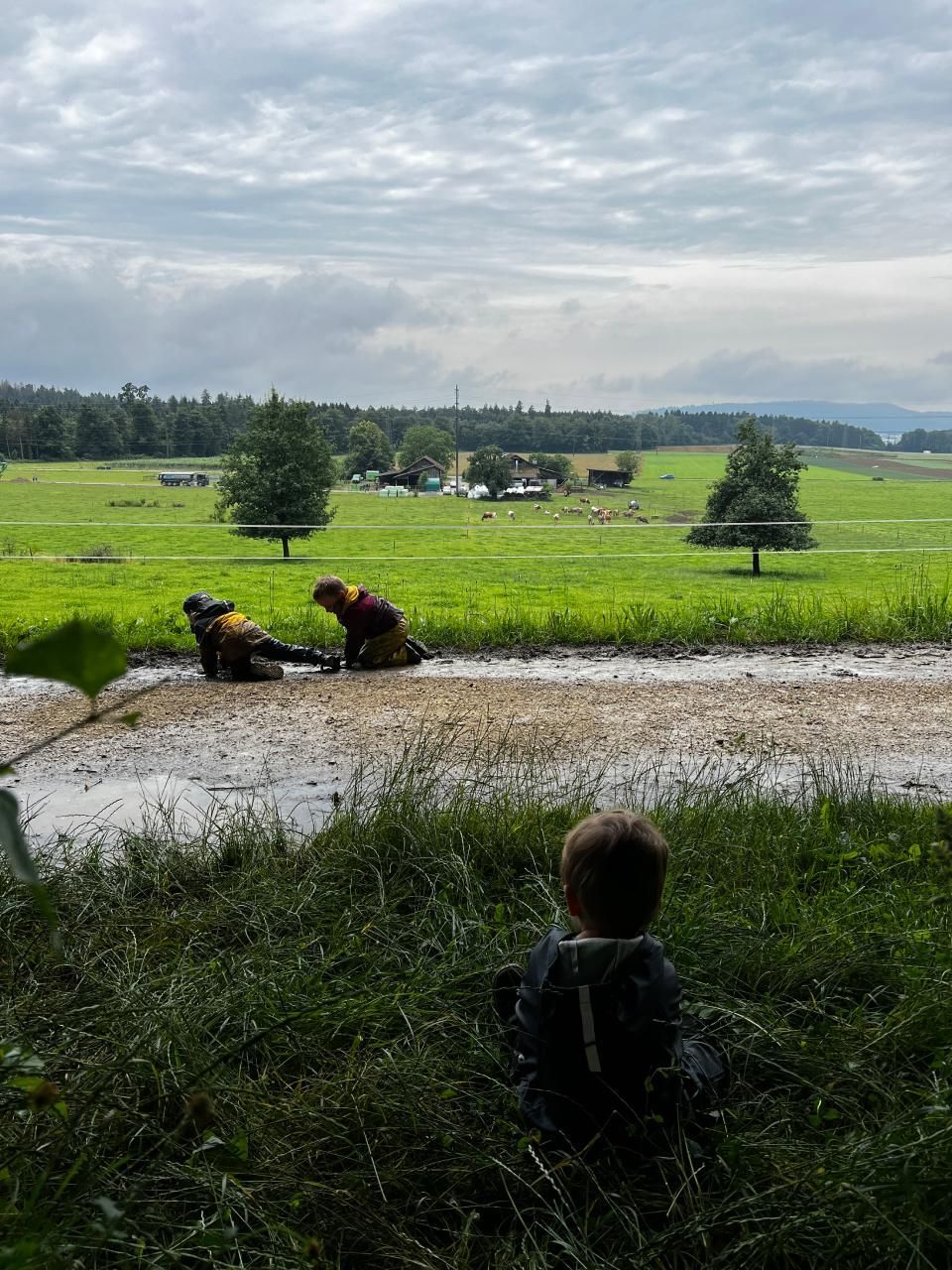 Ein Kind beobachtet zwei Personen in gelben Anzügen in einem schlammigen Gebiet, im Hintergrund ein grünes Feld. 