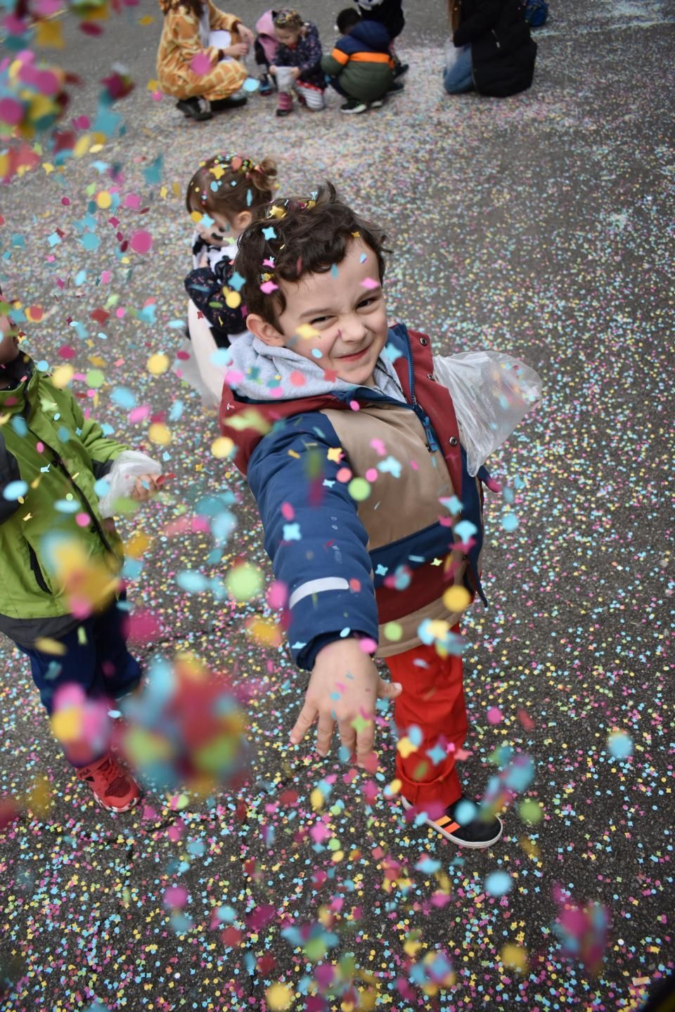 Ein lächelnder Junge streckt die Hand aus, Konfetti rieselt herab; im Hintergrund sind weitere Kinder zu sehen.