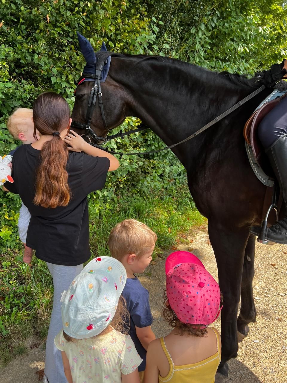 Eine Frau und Kinder streicheln ein schwarzes Pferd auf einem Pfad; das Pferd trägt Zaumzeug und Sattel.