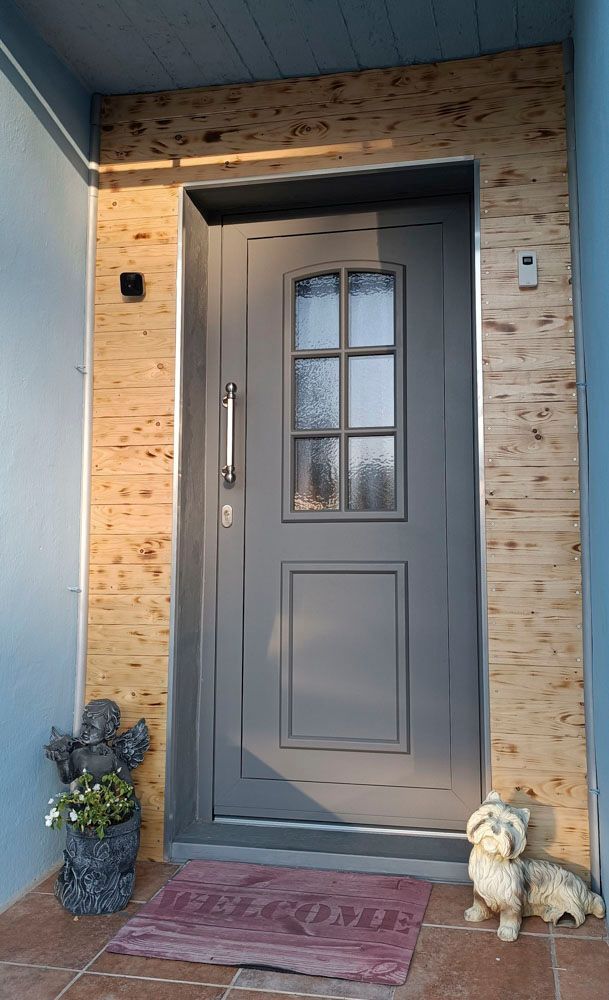Graue Haustür mit Glasscheiben und herbstlicher Dekoration, eingerahmt von hellem Holzpaneel und einer roten Fußmatte