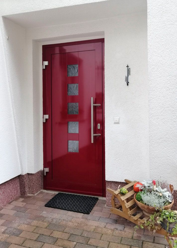 Rote Haustür mit kleinen Fenstern, silbernem Türgriff und Topfblumen vor einem gefliesten Eingangsbereich.
